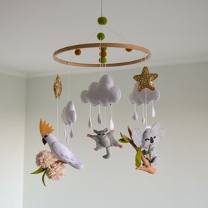 K&ouml;nnte beinhalten: Ein wei&szlig;es, graues und goldenes Filz-Babymobil mit einem Holzring. Das Mobile zeigt eine Wolke mit Regentropfen, einen Stern, einen Koala, einen Gleitbeutler und einen Kakadu.