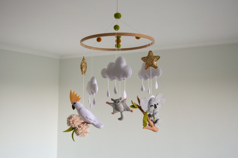 K&ouml;nnte beinhalten: Ein wei&szlig;es, graues und goldenes Filz-Babymobil mit einem Holzring. Das Mobile zeigt eine Wolke mit Regentropfen, einen Stern, einen Koala, einen Gleitbeutler und einen Kakadu.