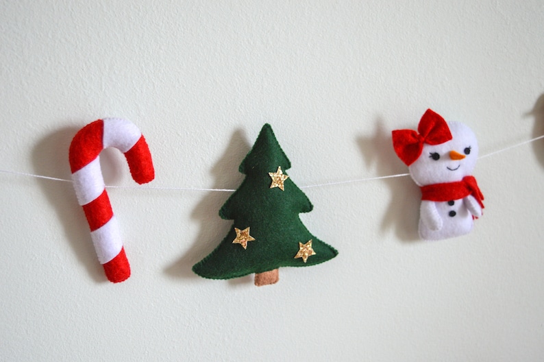 May include: A festive garland with a red and white candy cane, a green felt Christmas tree with gold glitter stars, and a white felt snowman with a red bow and scarf.