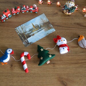 May include: A felt Christmas garland with a blue bird, a candy cane, a green Christmas tree, a snowman, a robin, and holly.