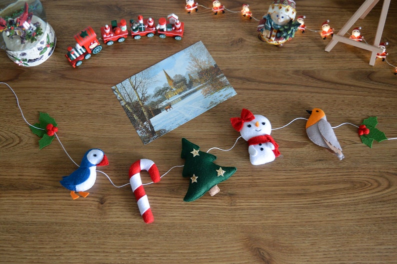 May include: A felt Christmas garland with a blue bird, a candy cane, a green Christmas tree, a snowman, a robin, and holly.