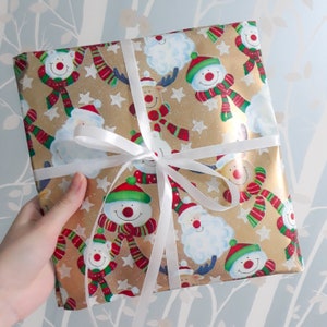 May include: A gold and white gift wrap with a festive Christmas design. The paper features a repeating pattern of snowmen, reindeer, and snowflakes. A white ribbon is tied around the package.