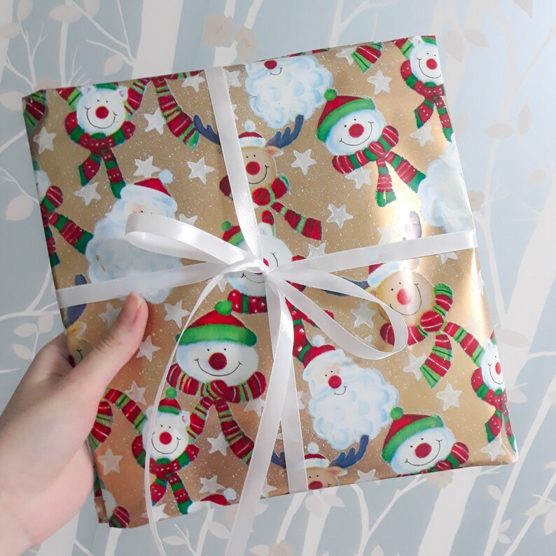 May include: A gold and white gift wrap with a festive Christmas design. The paper features a repeating pattern of snowmen, reindeer, and snowflakes. A white ribbon is tied around the package.