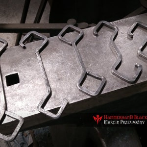 May include: Five metal hooks with a square shape at the top and a curved design. The hooks are arranged in a row on a metal surface. The hooks are made by Hammerhand Blacksmith Marcin Przewózmy.
