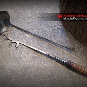 Op de afbeelding: Twee metalen gereedschappen, een lepel met een lange steel en een haak met een houten steel. De gereedschappen liggen op een grijs betonnen oppervlak. De tekst "Hammerhand Blacksmith Marcin Przewózmy" is zichtbaar in de rechterbovenhoek van de afbeelding.