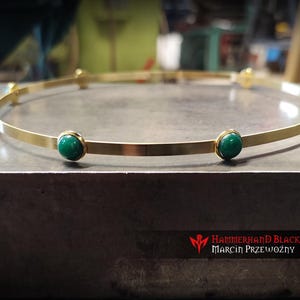 May include: A gold-colored crown with seven round, green gemstones evenly spaced around the band. The crown is displayed on a gray surface. The Hammerhand Blacksmith logo and name are visible in the bottom right corner.