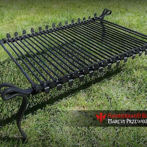 May include: Black metal grill grate with four legs. The grate is made of thick metal rods and has a decorative design on the legs. The grill is sitting on a green grassy surface.
