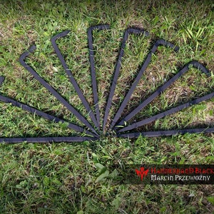 May include: Eight black metal stakes, each with a hooked end and a pointed tip, are fanned out on green grass. The stakes are arranged in a semi-circle. The image includes the text "Hammerhand Blacksmith Marcin Przewoźny".