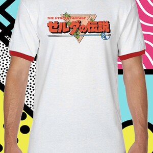 K&ouml;nnte beinhalten: Wei&szlig;es T-Shirt mit rotem Ringel, mit einem roten Dreieck mit dem Text "THE HYRULE FANTASY" und "ゼルダの伝説" in Japanisch.