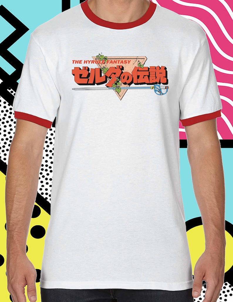 K&ouml;nnte beinhalten: Wei&szlig;es T-Shirt mit rotem Ringel, mit einem roten Dreieck mit dem Text "THE HYRULE FANTASY" und "ゼルダの伝説" in Japanisch.