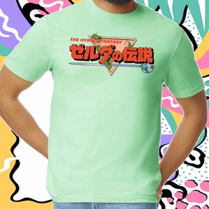 K&ouml;nnte beinhalten: Ein hellgr&uuml;nes T-Shirt mit einem Grafikdruck eines Schwertes und eines Dreiecks mit dem Text "THE HYRULE FANTASY" und "ゼルダの伝説" in Japanisch.