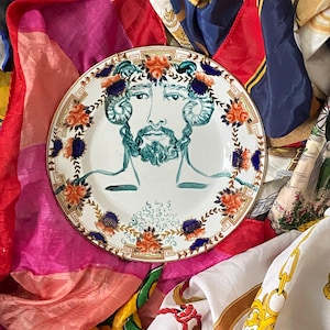 Op de afbeelding: Een decoratieve keramische plaat met een turquoise illustratie van een man met een baard en bloemmotieven. De plaat heeft een gouden rand en is omgeven door kleurrijke zijden sjaals.