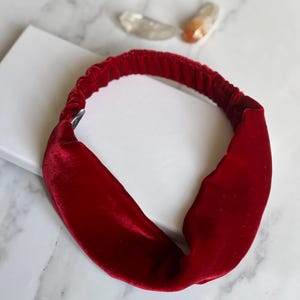 Op de afbeelding: Een dieprode fluwelen haarband met een gedraaid knoopontwerp. De haarband is gemaakt van een zacht, pluche materiaal en is waarschijnlijk voor mode of haarstyling. De achtergrond is een wit oppervlak met een marmerpatroon.