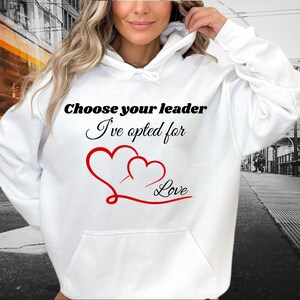 Peut inclure: Sweat-shirt à capuche blanc avec un motif noir qui dit "Choose your leader I've opted for Love" avec deux cœurs rouges.
