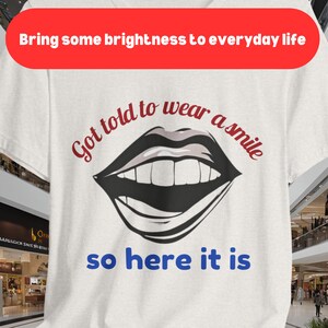 以下が含まれることがあります： 白いTシャツに、黒と白の笑顔の口のイラストと「Got told to wear a smile so here it is」というテキストが書かれています。