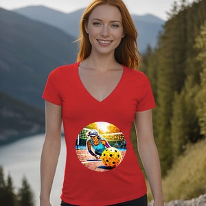 Peut inclure: T-shirt rouge à col en V avec un graphique circulaire d'une femme jouant au pickleball. Le graphique est coloré et présente un arrière-plan de coucher de soleil.