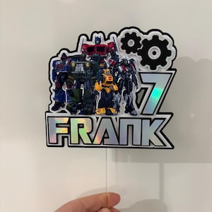 May include: A cake topper with a group of Transformers characters, the number 7, and the name "FRANK" in a holographic font. The design includes black and white accents and a clear stick for insertion.