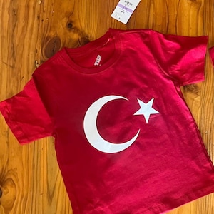 May include: Red t-shirt featuring a white crescent moon and star design. The shirt has the word "TILT" printed on the collar.