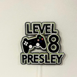 May include: A layered cake topper with the words "LEVEL 8 PRESLEY" in black and white, with a green outline. A black controller graphic is in the center. The number 8 is also in black and white. The topper is on a clear stick.