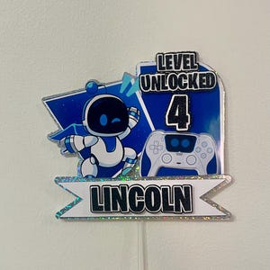 May include: A blue and white cake topper featuring a robot, a video game controller, and the text "Level Unlocked 4" and "Lincoln".