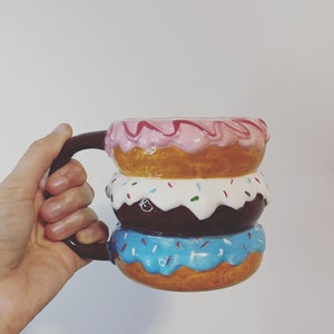 May include: A novelty ceramic mug designed as a stack of three donuts. The top donut has pink icing, the middle has white icing with sprinkles, and the bottom has blue icing. The mug has a brown handle.