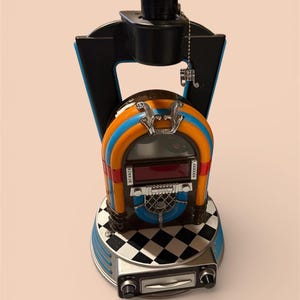 May include: A retro jukebox lamp with a black top and a colourful body featuring orange, blue, and red accents. The base has a checkerboard pattern and a silver-coloured control panel. The lamp has a pull chain.