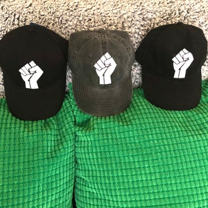 May include: Three black baseball caps with a white raised fist graphic on the front. The caps are on a green fuzzy surface.