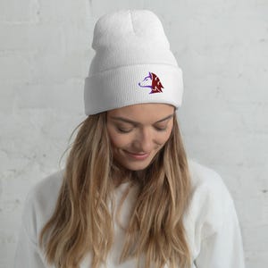 Puede incluir: Un gorro blanco con borde doblado. El gorro presenta un diseño de cabeza de lobo morado y rojo en la parte delantera. El gorro está hecho de un material de punto suave.