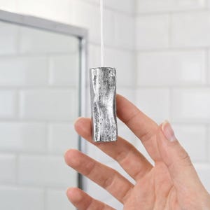 Peut inclure: Une tirette de lumière cylindrique argentée avec une surface texturée et irrégulière, fixée à un cordon blanc. L'objet présente une finition métallique avec un aspect brut et organique, orienté verticalement et suspendu par une fine ficelle blanche.
