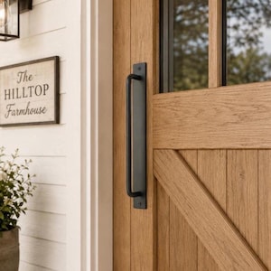 May include: A matte black metal door handle mounted vertically on a light brown wooden door with a diagonal brace. To the left, a rectangular sign with a dark frame displays the text The HILLTOP Farmhouse in black lettering on a cream background.