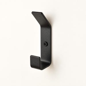 May include: A matte black metal wall hook featuring a J-shaped design with an upward-angled top prong and a lower hook. The piece is secured to the wall with a single black Phillips head screw positioned in the centre of the vertical mounting plate.