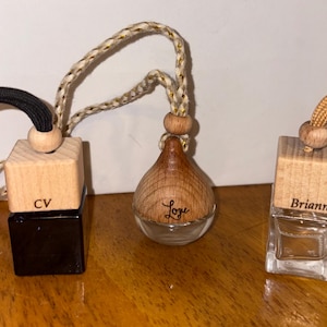 Puede incluir: Tres frascos colgantes para ambientador de coche con tapas de madera y cordones. El frasco izquierdo es negro con el texto CV, el central es redondo con el texto Love y el derecho es de cristal transparente con el texto Briann en la tapa.