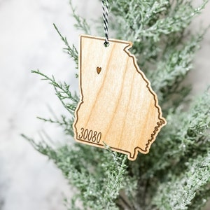 May include: Wooden ornament in the shape of the state of Georgia, with a small heart and the zip code 30080 etched into the wood. The ornament hangs from a black and white string against a backdrop of a green, frosted tree.