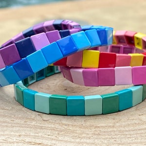 May include: A stack of colorful square tile bracelets in shades of blue, green, pink, red, and yellow.