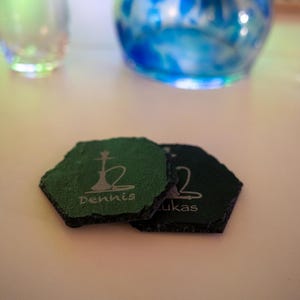 Könnte beinhalten: Zwei grüne und schwarze Schieferuntersetzer mit einem Wasserpfeifen-Design und den eingravierten Namen "Dennis" und "Lukas".