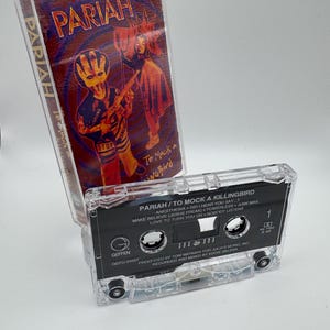 May include: A clear cassette tape with the album title "Pariah / To Mock a Killingbird" is displayed. The cassette case has a red and purple cover with a graphic of a figure holding a guitar. The cassette tape is on a white surface.