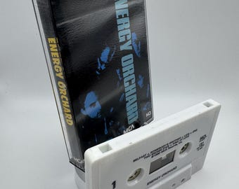 May include: A vintage audio cassette tape with the text "ENERGY ORCHARD" in blue and yellow. The cassette is white with the text "ENERGY ORCHARD" and song titles. The cassette case is black.