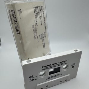 May include: A white Dharma Bums "Welcome" cassette tape with the tracklist visible. The cassette case is open, revealing the tape's label and the album title. The cassette tape is a vintage music collectible.