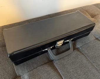 May include: A black rectangular case with a handle and a silver clasp. The case is sitting on a gray, textured surface. The case appears to be made of a shiny material.