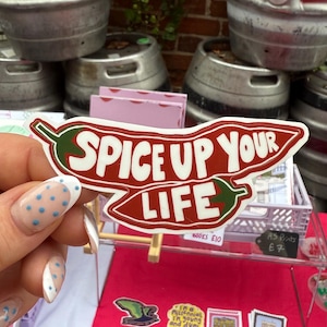 May include: A sticker in the shape of a chilli pepper, featuring the text "SPICE UP YOUR LIFE". The sticker is red and white, and is held in front of a market stall with other items.