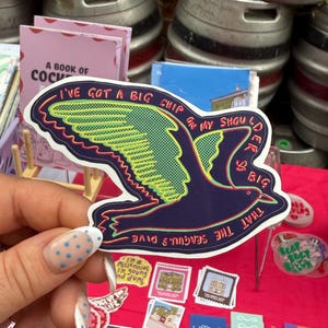 May include: A navy blue seagull sticker with green and yellow accents, and the text "I'VE GOT A BIG CHIP ON MY SHOULDER SO BIG THAT THE SEAGULLS DIVE." The sticker is held up against a red surface with other stickers and books visible in the background.