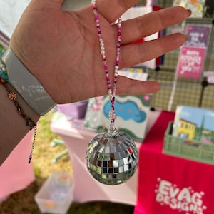 Può includere: Un ornamento a sfera da discoteca argentata con una catena di perline, tenuto in mano. La catena presenta perline bianche, rosa e viola. La sfera da discoteca è composta da piccoli quadrati a specchio. Lo sfondo include un tavolo rosa e biglietti di auguri.