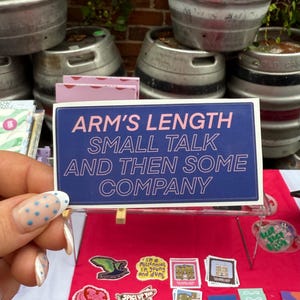 Op de afbeelding: Een blauw bord met de tekst "ARM'S LENGTH SMALL TALK AND THEN SOME COMPANY" wordt getoond voor een marktkraam. Verschillende stickers en andere items zijn zichtbaar, met bierfusten op de achtergrond.