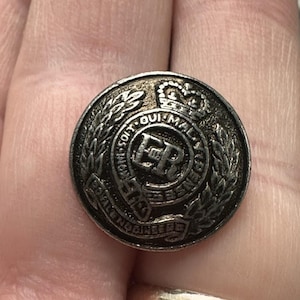 Puede incluir: Un botón antiguo detallado en tono plateado con una corona, una corona de laurel y texto. El botón presenta las letras "ER" en el centro, rodeadas por las palabras "SOIT QUI MAL Y PENSE" y "ROYAL ENGINEERS".