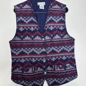 May include: A patterned vest in shades of burgundy, gray, and teal, with a navy blue lining and a V-neck. The vest has a button-up front and two front pockets. The vest is hanging on a black hanger.