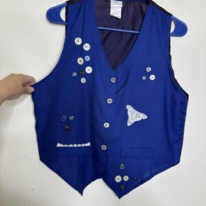 May include: A royal blue vest with a V-neck, adorned with various buttons and a white lace appliqué. The vest has a dark purple lining and is hanging on a blue hanger.