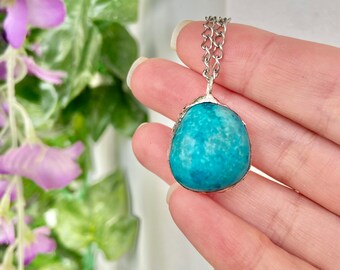 May include: A silver chain necklace with a large, round, turquoise stone pendant. The stone has a textured surface and is set in a silver bezel. The necklace is shown against a backdrop of green leaves and purple flowers.