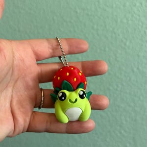 May include: A small keychain featuring a light green frog with a white belly and large black eyes. The frog has a bright red strawberry with yellow seeds resting on its head, surrounded by small green leaves. A silver metal ball chain is attached.