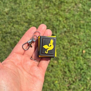May include: A small, rectangular keychain with a black and yellow design. The keychain features a yellow silhouette of a figure on a black background. The keychain is attached to a silver-colored clasp.