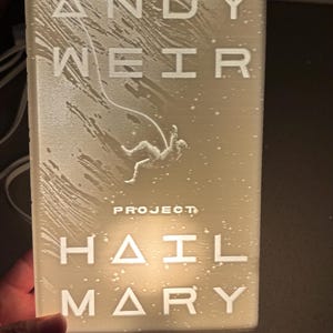 May include: A rectangular, illuminated light featuring the book cover of "Project Hail Mary" by Andy Weir. The design includes the author's name, book title, and an illustration of a figure in space. The light is held in a person's hand.